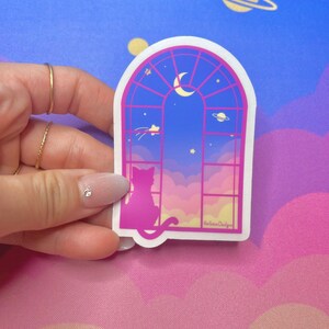 May include: A pink and blue sticker with a cat silhouette looking out a window at a crescent moon and stars. The window is framed in pink and has a gold 'HallieRae Designs' logo in the bottom right corner.