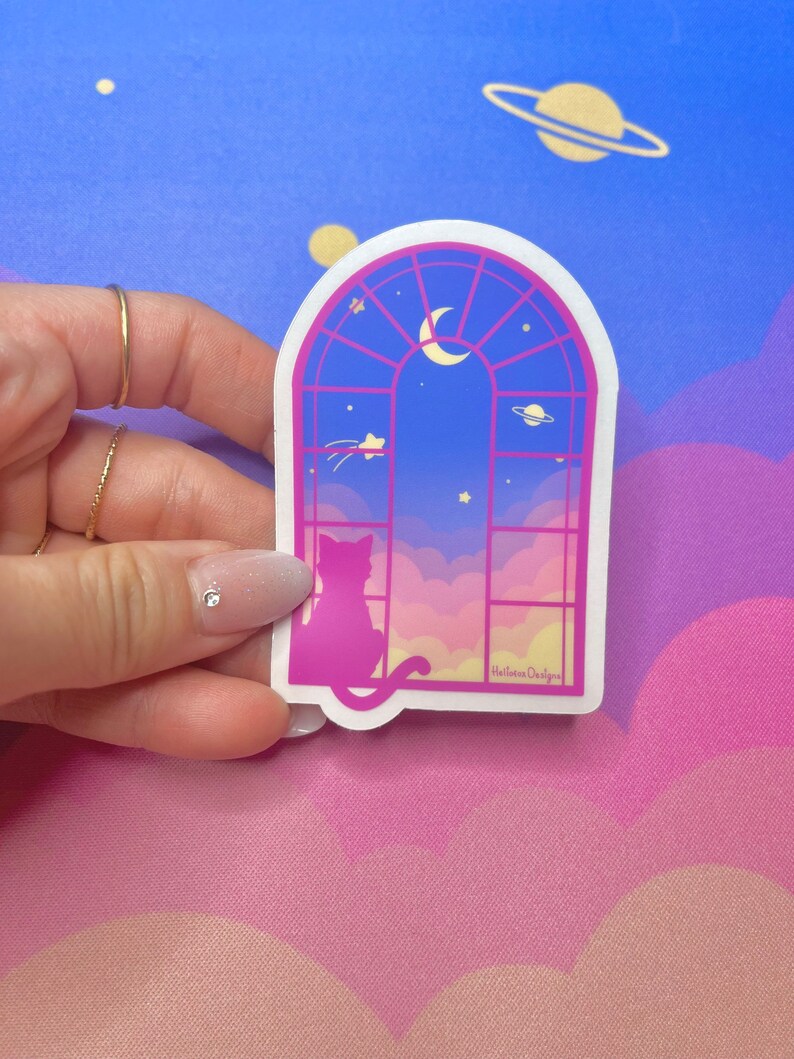 May include: A pink and blue sticker with a cat silhouette looking out a window at a crescent moon and stars. The window is framed in pink and has a gold 'HallieRae Designs' logo in the bottom right corner.