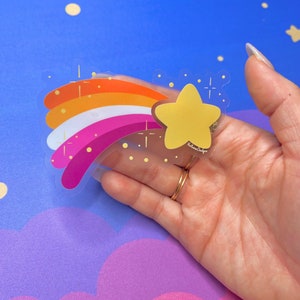 May include: A clear sticker with a rainbow design in the colors of the lesbian pride flag. A yellow star with a gold outline is at the end of the rainbow. The sticker has a white background and is on a blue surface.