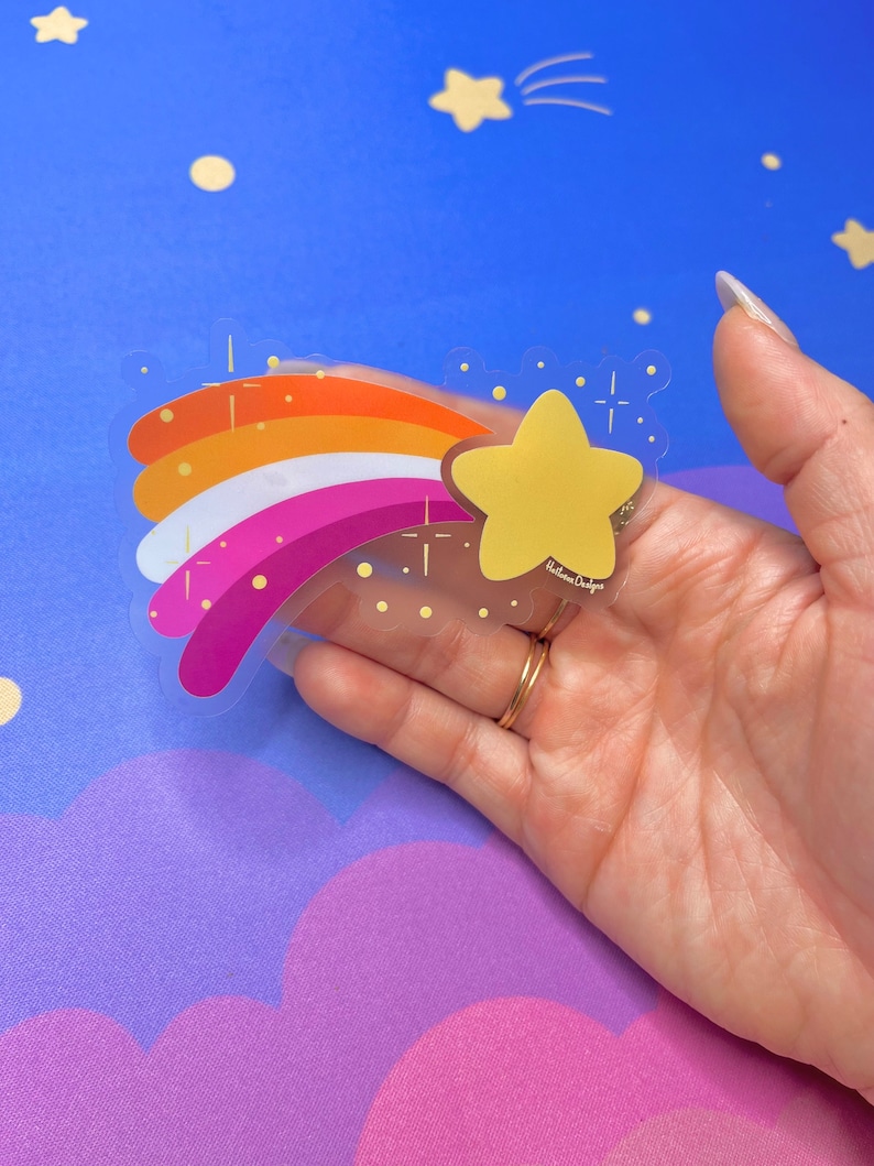 May include: A clear sticker with a rainbow design in the colors of the lesbian pride flag. A yellow star with a gold outline is at the end of the rainbow. The sticker has a white background and is on a blue surface.