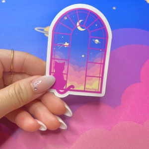 May include: A sticker featuring a pink and blue window with a crescent moon and stars. A silhouette of a cat sits in the window looking out at a pink and yellow sky with clouds. The sticker has the text "Hellolove Designs" at the bottom.