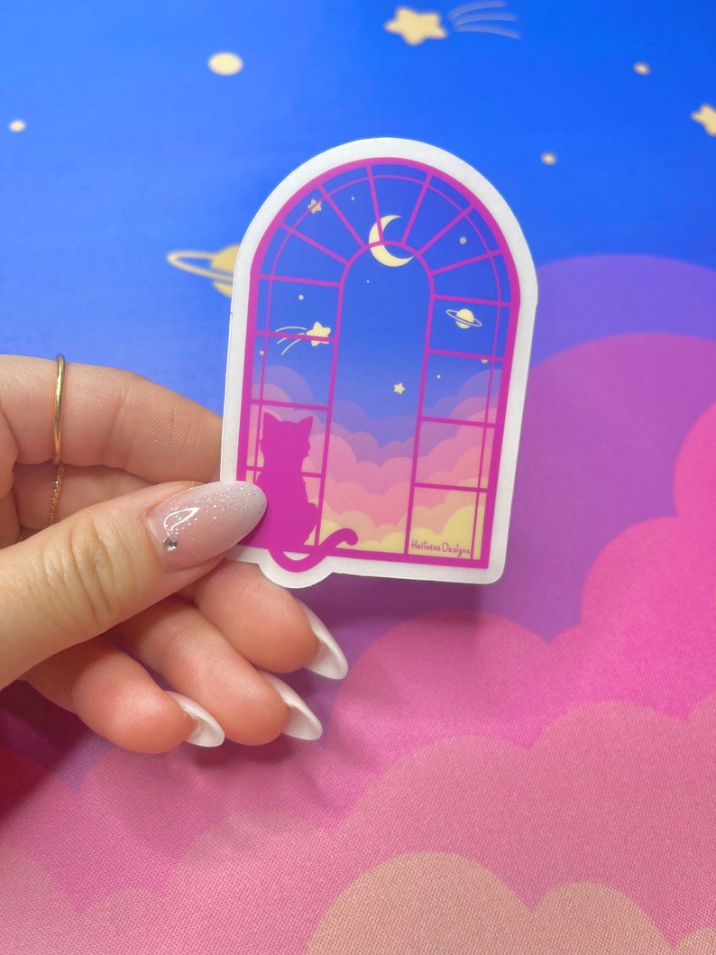 May include: A sticker featuring a pink and blue window with a crescent moon and stars. A silhouette of a cat sits in the window looking out at a pink and yellow sky with clouds. The sticker has the text "Hellolove Designs" at the bottom.
