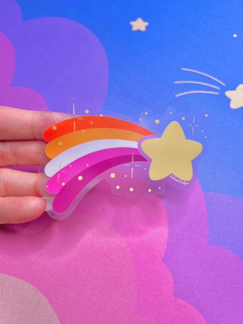 May include: A clear sticker with a rainbow design in the colors of the lesbian pride flag. The rainbow is shooting up towards a yellow star with a glitter effect. The sticker has the text "Rainbow Designs" in the bottom right corner.