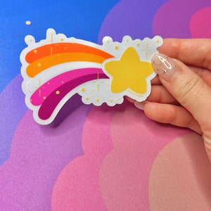 May include: A rainbow-colored shooting star sticker with a yellow star at the end. The sticker has a white background and gold glitter accents.