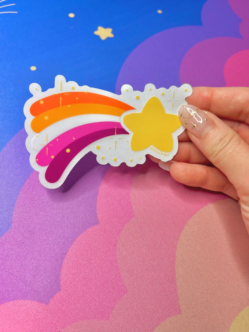 May include: A rainbow-colored shooting star sticker with a yellow star at the end. The sticker has a white background and gold glitter accents.