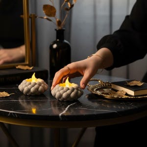 Op de afbeelding: Twee grijze, getextureerde kaarshouders met gele LED-vlammen op een zwarte marmeren tafel. Een hand reikt naar een van de kaarshouders. Een goudkleurig dienblad en spiegel staan op de achtergrond.