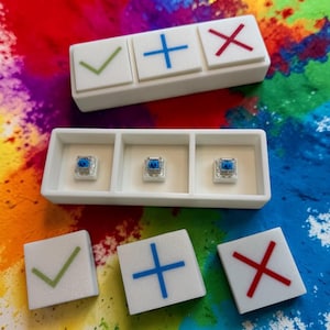 Op de afbeelding: Witte vierkante toetsdoppen met een groene vink, een blauw plusteken en een rood kruis symbool. De afbeelding toont ook drie toetsdoppen met een transparante, blauw getinte schakelaar. De achtergrond is een kleurrijke verfspat.