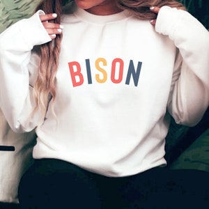 May include: White sweatshirt with the word "BISON" printed in red, yellow, blue, and white letters.