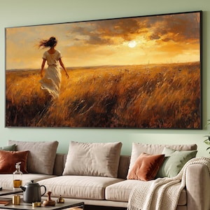 Op de afbeelding: Een rechthoekige wandkunstprint in een zwarte lijst met een persoon in een witte jurk die bij zonsondergang door een goudkleurig tarweveld loopt. De lucht is gevuld met warme oranje en gele wolken en het hoge gras is in amberkleurige tinten.