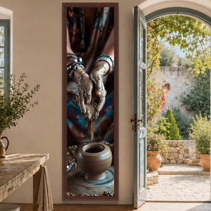 May include: A tall, rectangular wall art piece featuring a dark brown frame. The image depicts a pair of hands covered in wet, gray clay, shaping a small, smooth, light brown ceramic pot on a spinning pottery wheel, with blurred colorful clothing.