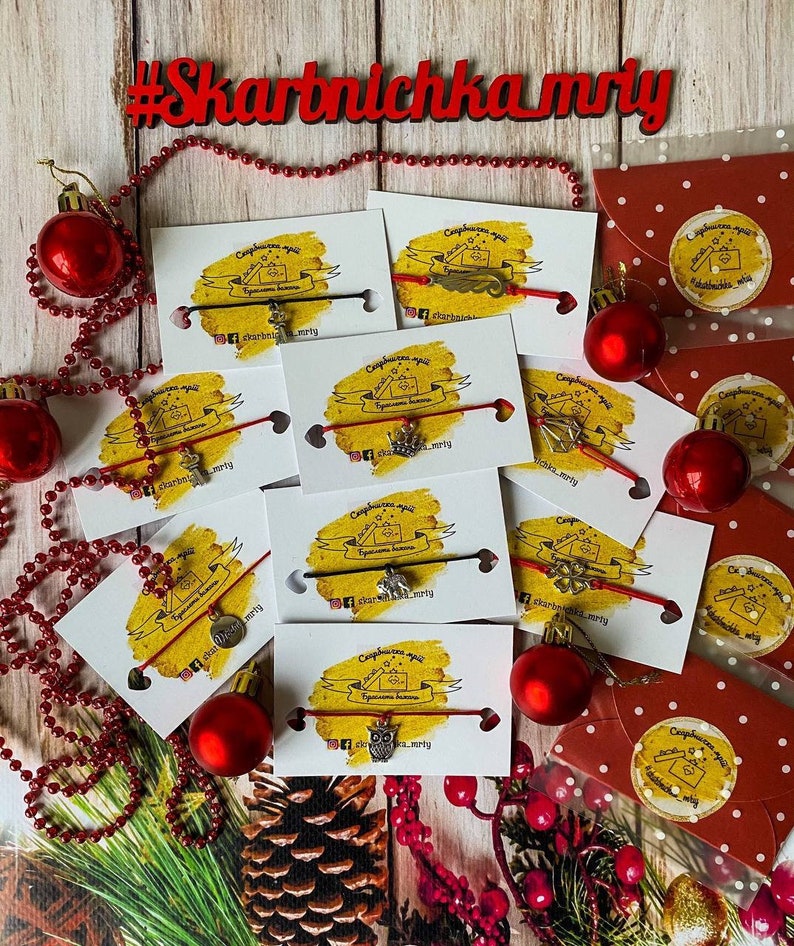 May include: A collection of red string bracelets with various charms, including a house, an owl, a star, and a key. The bracelets are displayed on white cards with a yellow background and the text "Скарбничка мрій" which translates to "Treasure Box of Dreams".