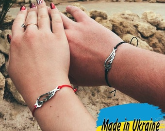 May include: Two matching bracelets with a silver wing charm on red and black string. The bracelets are worn on the wrists of two people. The text "Made in Ukraine" is visible in the image.