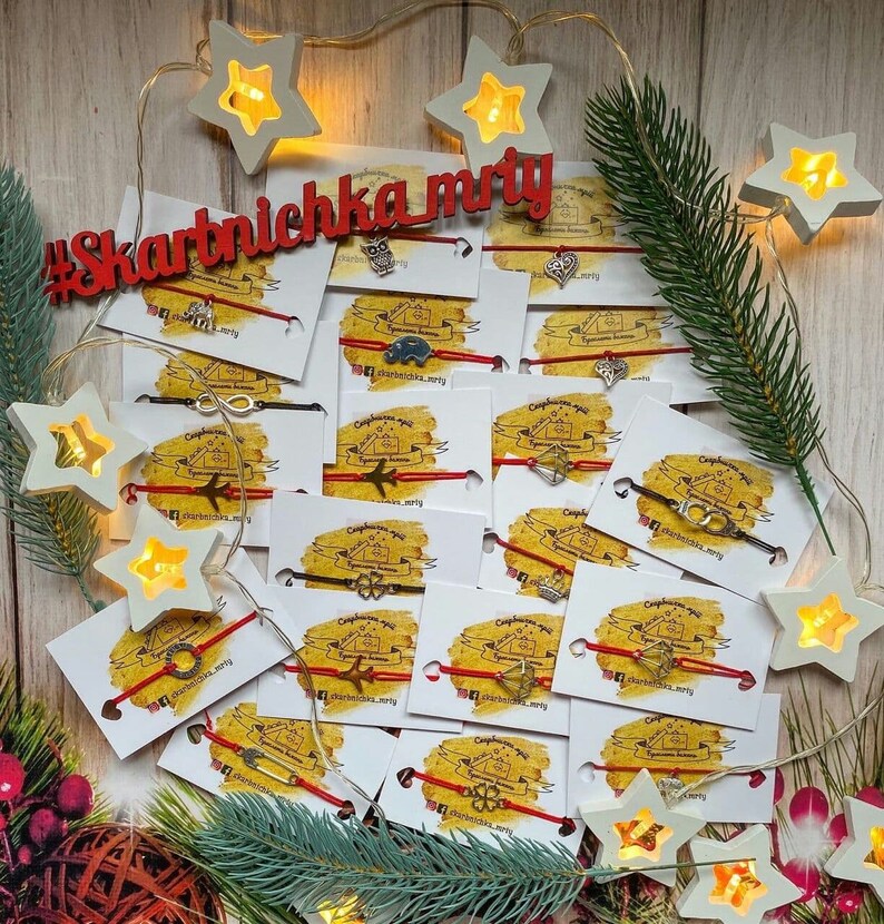 May include: A collection of red string bracelets with various silver charms, including a house, an airplane, an infinity symbol, a four-leaf clover, and a compass. The bracelets are displayed on white cards with the text "#skarbniczka_mry" and a website address.