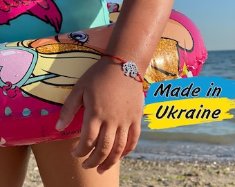 May include: A red string bracelet with a silver tree of life charm. The bracelet is worn on the wrist of a child wearing a blue swimsuit and a pink flamingo float. The background is a sandy beach with blue water. The text "Made in Ukraine" is written in blue and yellow on a white background.