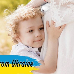 Może przedstawiać: Małe dziecko z kręconymi blond włosami przytula osobę ubraną w białą sukienkę. Dziecko patrzy w obiektyw z uśmiechem. Obraz zawiera tekst "From Ukraine" w kolorze żółtym i niebieskim, kolorach flagi ukraińskiej.