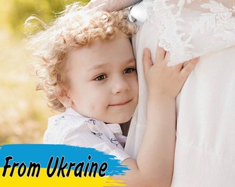 May include: A young child with curly blonde hair hugs a person wearing a white dress. The child is looking at the camera with a smile. The image has the text "From Ukraine" in yellow and blue, the colors of the Ukrainian flag.