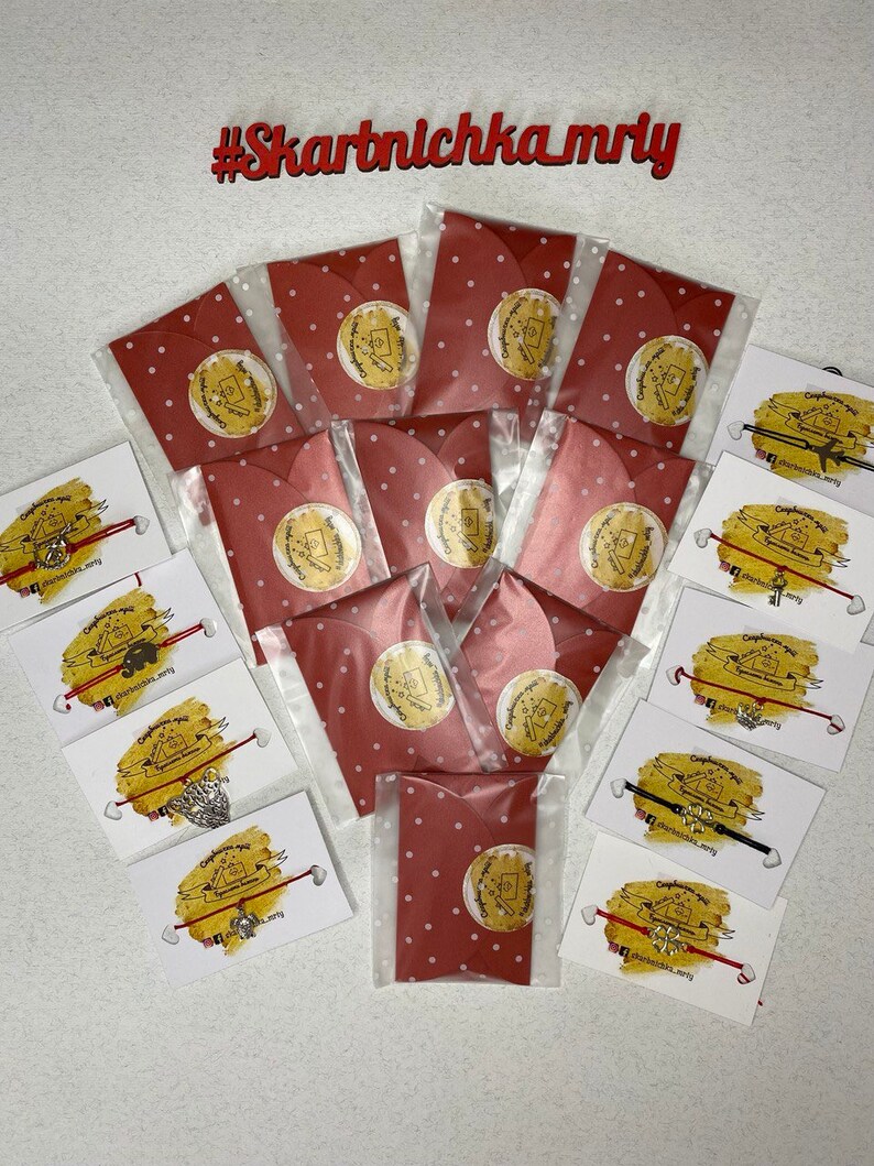 May include: A collection of red and white polka dot envelopes with a small white circle with a design in the center. The envelopes are arranged in a semi-circle around a collection of red and black string bracelets with silver charms. The bracelets are on white cards with the text "#Skartnlchka_mry" on them.