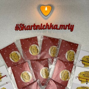 May include: Red envelopes with white polka dots and gold foil stickers that say "Congratulations!"  The envelopes are arranged in a circle around a lit heart-shaped candle. The hashtag #Skartnlchka_mriy is written in red letters on a white background.