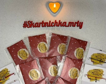 May include: Red envelopes with white polka dots and gold foil stickers that say "Congratulations!"  The envelopes are arranged in a circle around a lit heart-shaped candle. The hashtag #Skartnlchka_mriy is written in red letters on a white background.