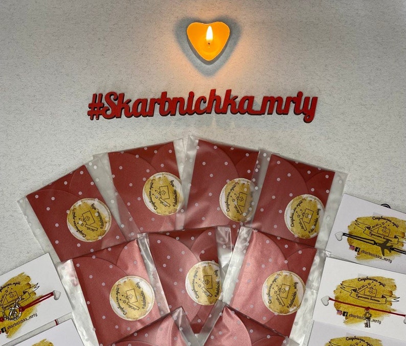 May include: Red envelopes with white polka dots and gold foil stickers that say "Congratulations!"  The envelopes are arranged in a circle around a lit heart-shaped candle. The hashtag #Skartnlchka_mriy is written in red letters on a white background.