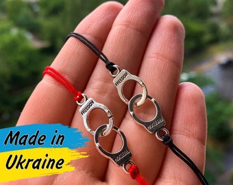 May include: Two bracelets with silver handcuff charms. The bracelets are made with red and black string. The charms have the word "FREEDOM" engraved on them. The bracelets are on a person's wrist. The image has the text "Made in Ukraine" on it.