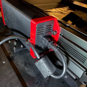 May include: A close-up of a black power supply unit with a red housing and a black power cord. The power supply unit is mounted to a metal surface with screws.