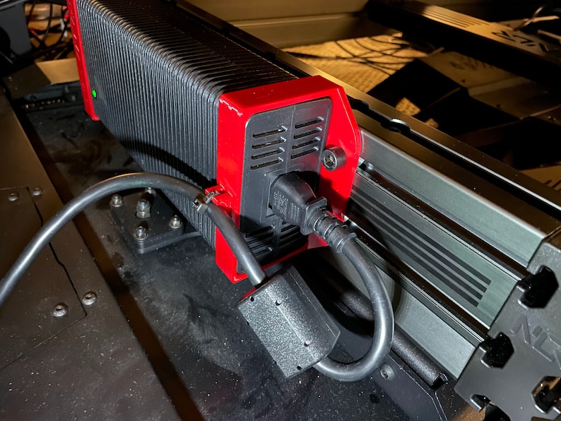 May include: A close-up of a black power supply unit with a red housing and a black power cord. The power supply unit is mounted to a metal surface with screws.