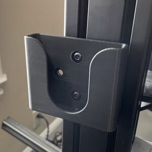 May include: A black plastic wall mount with a curved opening for holding a small object. The mount has four screw holes.