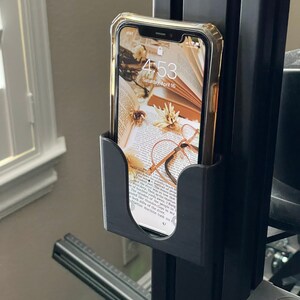 May include: A black phone holder attached to a black metal pole. A clear phone case with a floral wallpaper is in the holder. The phone screen displays the time 4:53 and the date Saturday, April 16.