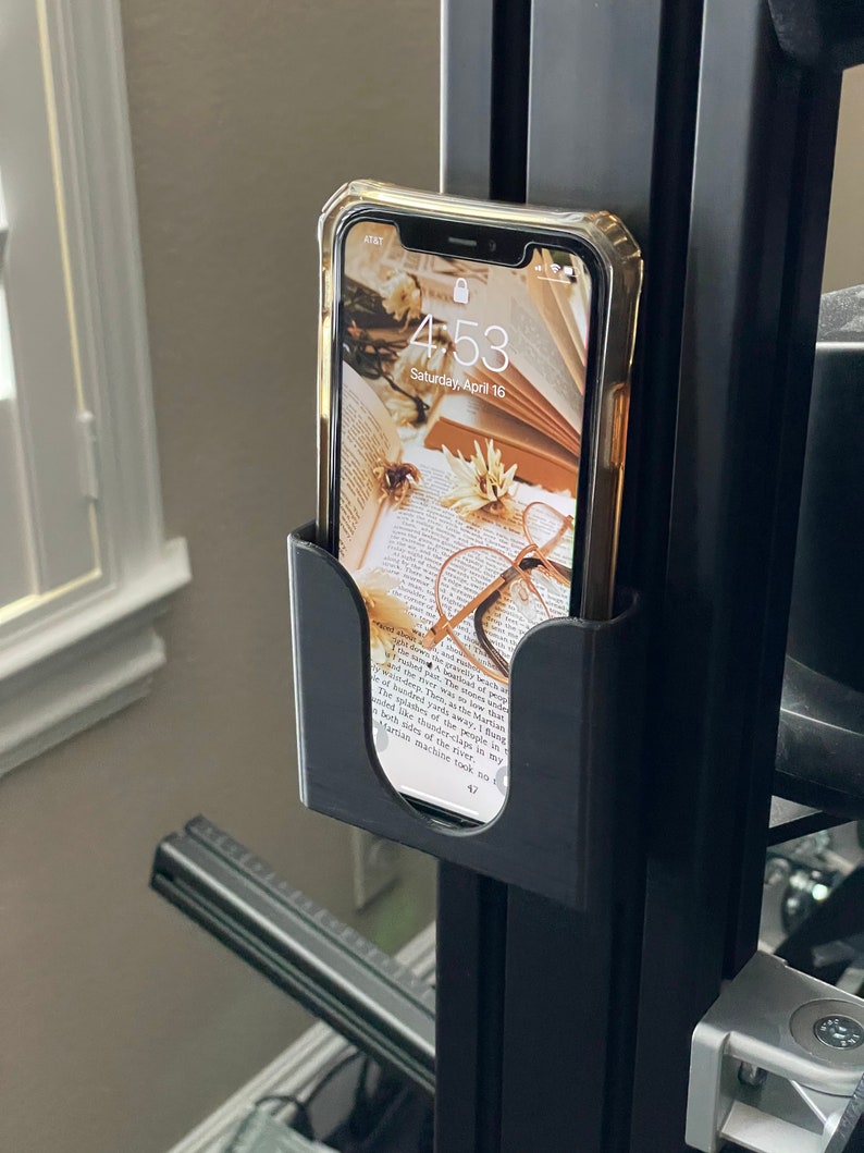 May include: A black phone holder attached to a black metal pole. A clear phone case with a floral wallpaper is in the holder. The phone screen displays the time 4:53 and the date Saturday, April 16.