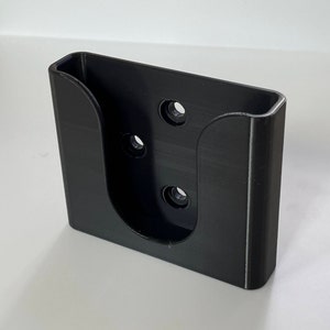May include: Black plastic wall mount with four holes for mounting screws. The mount has a U-shaped opening for holding an object.