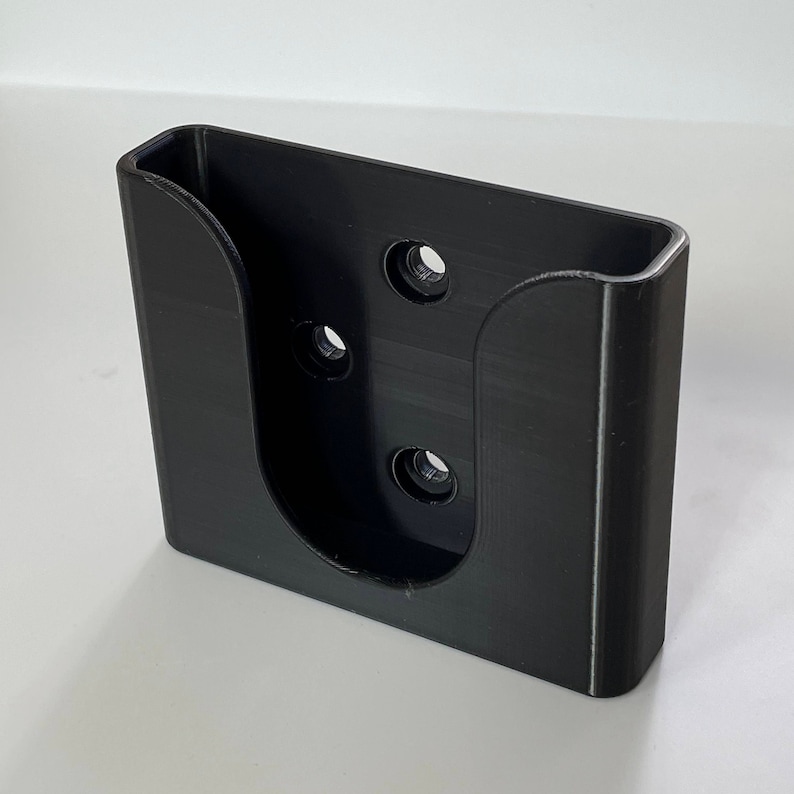 May include: Black plastic wall mount with four holes for mounting screws. The mount has a U-shaped opening for holding an object.
