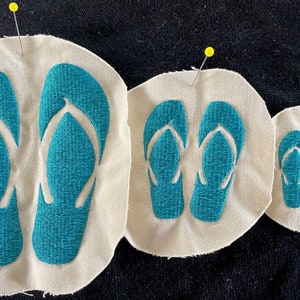 Op de afbeelding: Drie stoffen patches met geborduurde turquoise slippers. De patches zijn van verschillende grootte en zijn vastgemaakt aan een donkere stoffen achtergrond.