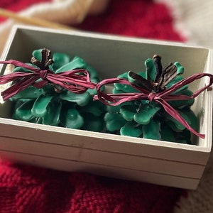 May include: Two green pine cone shaped candles with burgundy ribbon bows in a white wooden box.