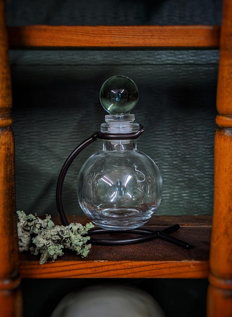 Puede incluir: Una botella de vidrio transparente con cuerpo redondo y tap&oacute;n de vidrio. La botella est&aacute; sobre un soporte de metal negro con asa curva. La botella est&aacute; sobre un estante de madera.