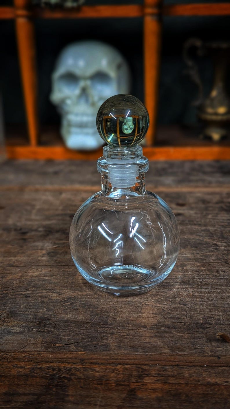 Puede incluir: Una botella de vidrio transparente con cuerpo redondo y un tap&oacute;n de vidrio. El tap&oacute;n es una esfera con una superficie texturizada. La botella est&aacute; sobre una superficie de madera.