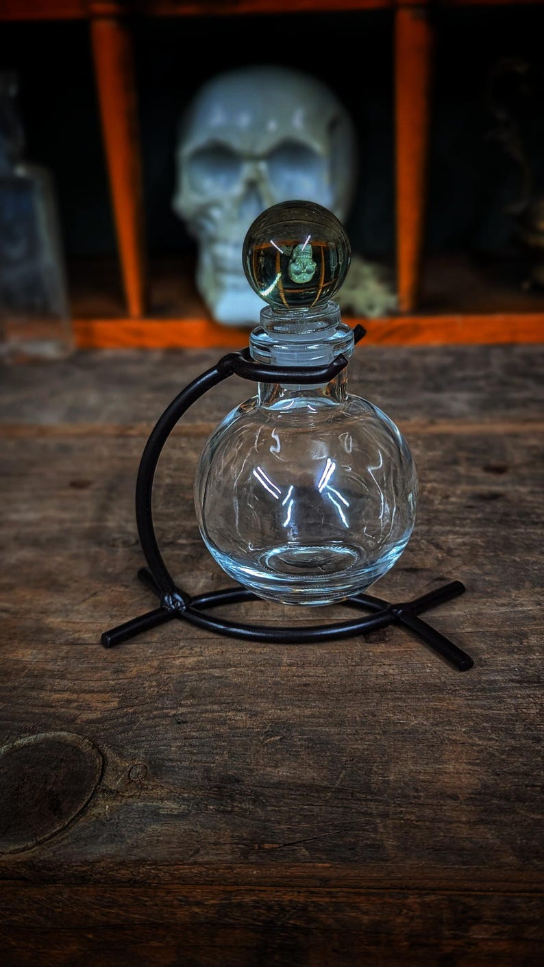 Puede incluir: Una botella de vidrio transparente con cuerpo redondo y tap&oacute;n de vidrio. La botella est&aacute; sobre un soporte de metal negro con base curva.