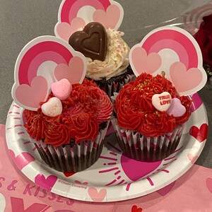 K&ouml;nnte beinhalten: Drei Schokoladen-Cupcakes, verziert mit rotem und wei&szlig;em Frosting. Die Cupcakes sind mit regenbogenf&ouml;rmigen Papierschnitten und herzf&ouml;rmigen S&uuml;&szlig;igkeiten verziert. Die Cupcakes befinden sich auf einem wei&szlig;en Pappteller mit einem rosa-wei&szlig;en XOXO-Design.