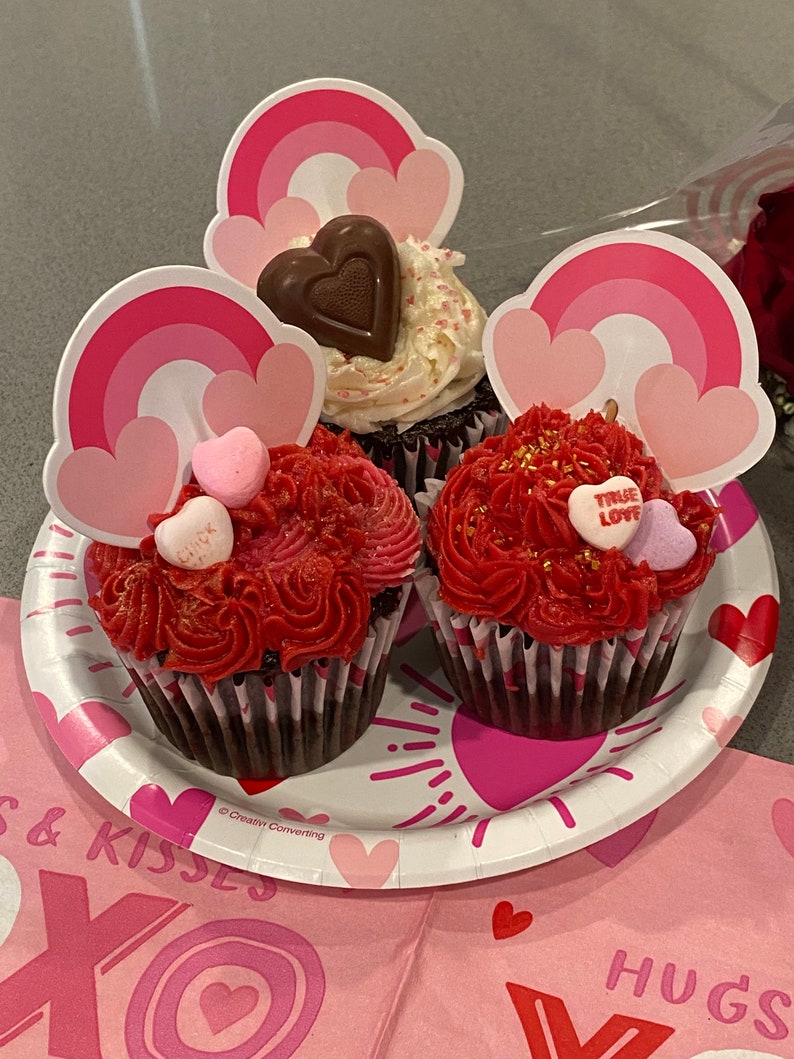 K&ouml;nnte beinhalten: Drei Schokoladen-Cupcakes, verziert mit rotem und wei&szlig;em Frosting. Die Cupcakes sind mit regenbogenf&ouml;rmigen Papierschnitten und herzf&ouml;rmigen S&uuml;&szlig;igkeiten verziert. Die Cupcakes befinden sich auf einem wei&szlig;en Pappteller mit einem rosa-wei&szlig;en XOXO-Design.