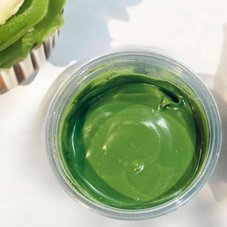 Op de afbeelding: Een close-up van een doorzichtige plastic container gevuld met felgroene glazuur. De glazuur heeft een gladde, glanzende textuur. Een deel van een cupcake met groene glazuur is linksboven zichtbaar.