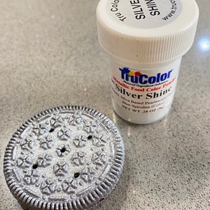 May include: A silver-colored, decorated cookie next to a container of TruColor Silver Shine metallic food color powder. The container has a white lid with the words "Silver Shine" printed on it. The cookie has a detailed pattern.