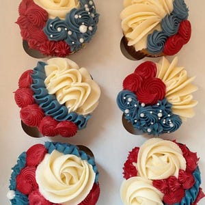 May include: Six cupcakes with white, red, and blue buttercream frosting, arranged in a box. The cupcakes are decorated with buttercream roses and swirls, with white pearl sprinkles. The text "@cottage_confections_bysara" is at the bottom.