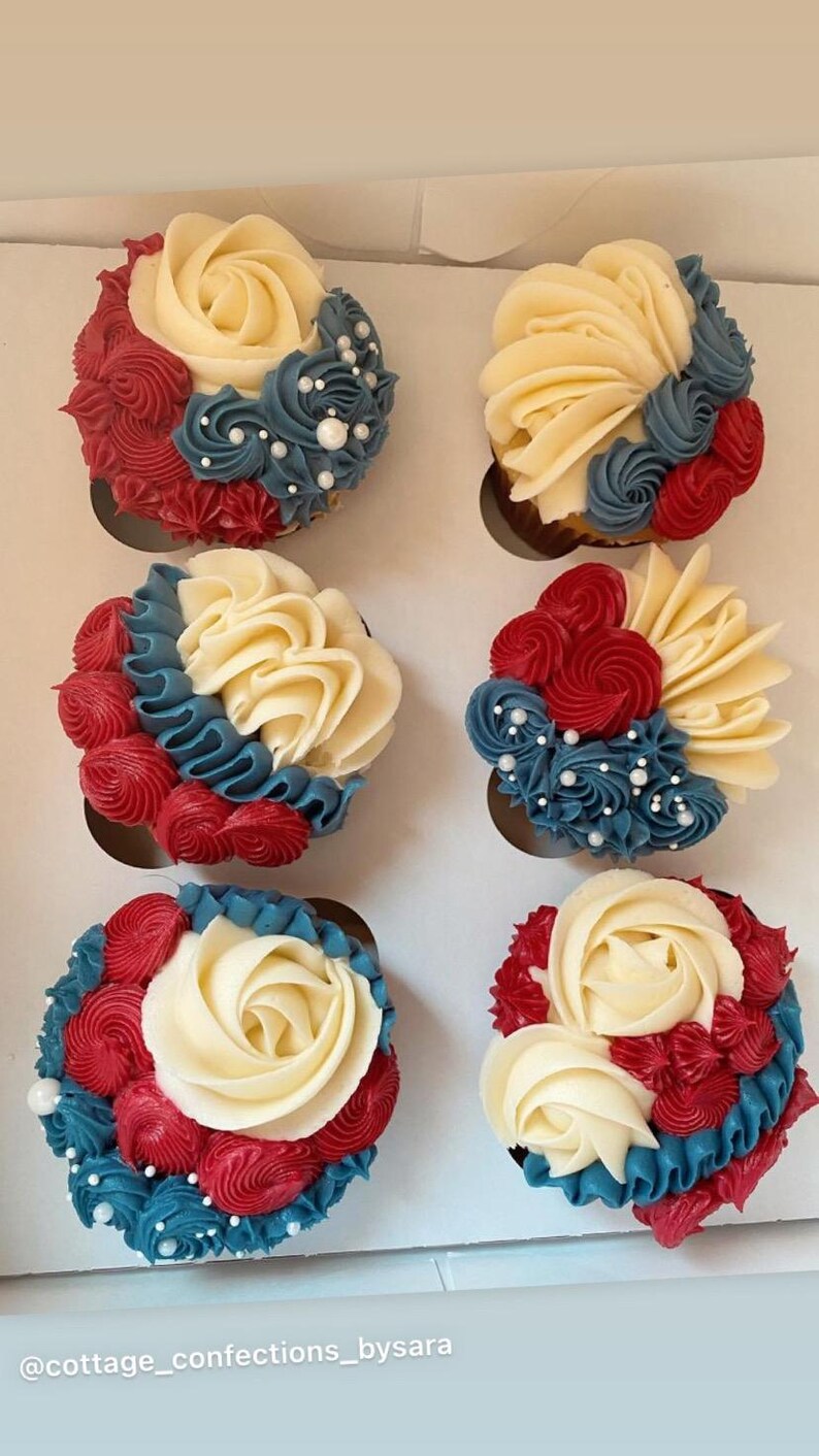 May include: Six cupcakes with white, red, and blue buttercream frosting, arranged in a box. The cupcakes are decorated with buttercream roses and swirls, with white pearl sprinkles. The text "@cottage_confections_bysara" is at the bottom.