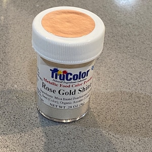 May include: A small jar of TruColor Rose Gold Shine metallic food color powder. The jar has a white lid with a rose gold colored top. The label lists ingredients and the net weight of .28 oz (8g).