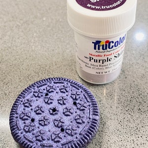 May include: A small jar of purple food coloring with the words "TruColor Purple Shine" on the label. The jar is next to a purple cookie with a star-shaped design.