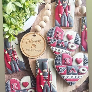 May include: A set of decorated sugar cookies featuring a red and black toy soldier design and a heart-shaped cookie with the text "I FAO" in black and red lettering. The cookies are on a wooden surface with a wooden bead garland.