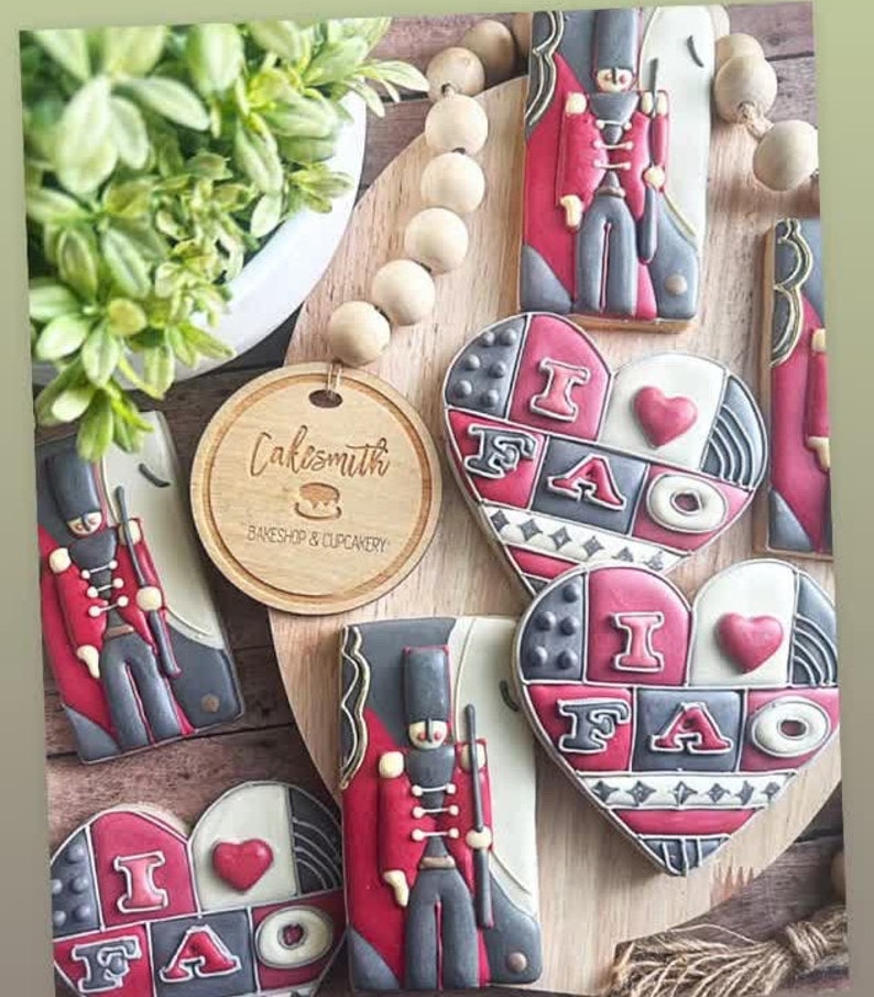 May include: A set of decorated sugar cookies featuring a red and black toy soldier design and a heart-shaped cookie with the text "I FAO" in black and red lettering. The cookies are on a wooden surface with a wooden bead garland.