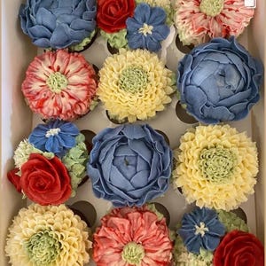 May include: A box of a dozen cupcakes, each topped with buttercream flowers in shades of blue, red, yellow, and pink, with green detailing. The floral designs include roses and other blooms. The cupcakes are arranged in a grid.
