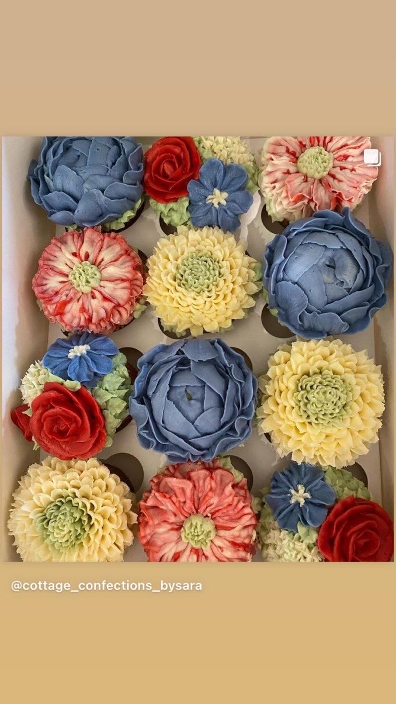 May include: A box of a dozen cupcakes, each topped with buttercream flowers in shades of blue, red, yellow, and pink, with green detailing. The floral designs include roses and other blooms. The cupcakes are arranged in a grid.