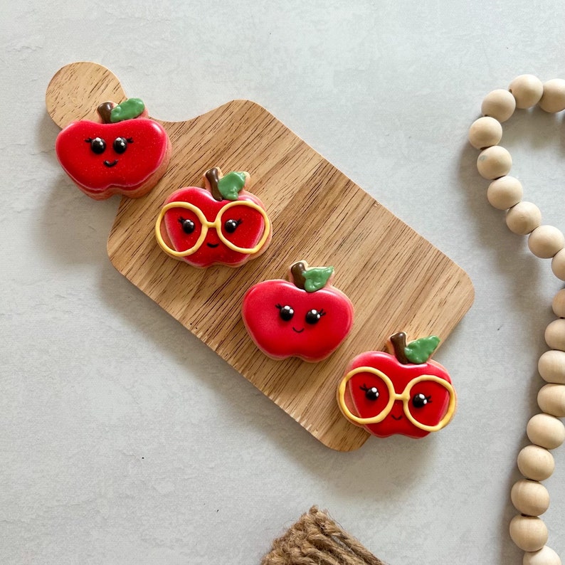 May include: Four red apple-shaped cookies decorated with white eyes, a smiling mouth, and green stems. Two of the cookies have yellow glasses.